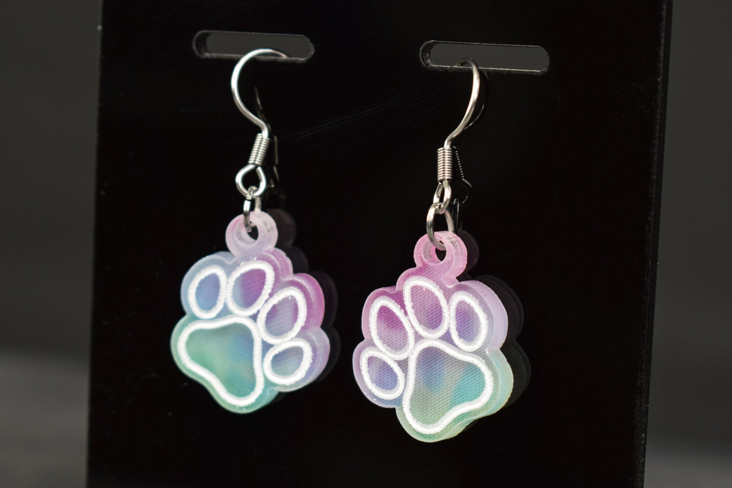 A pair of multicolored earrings with a paw print design and a cool hue, displayed on a black stand