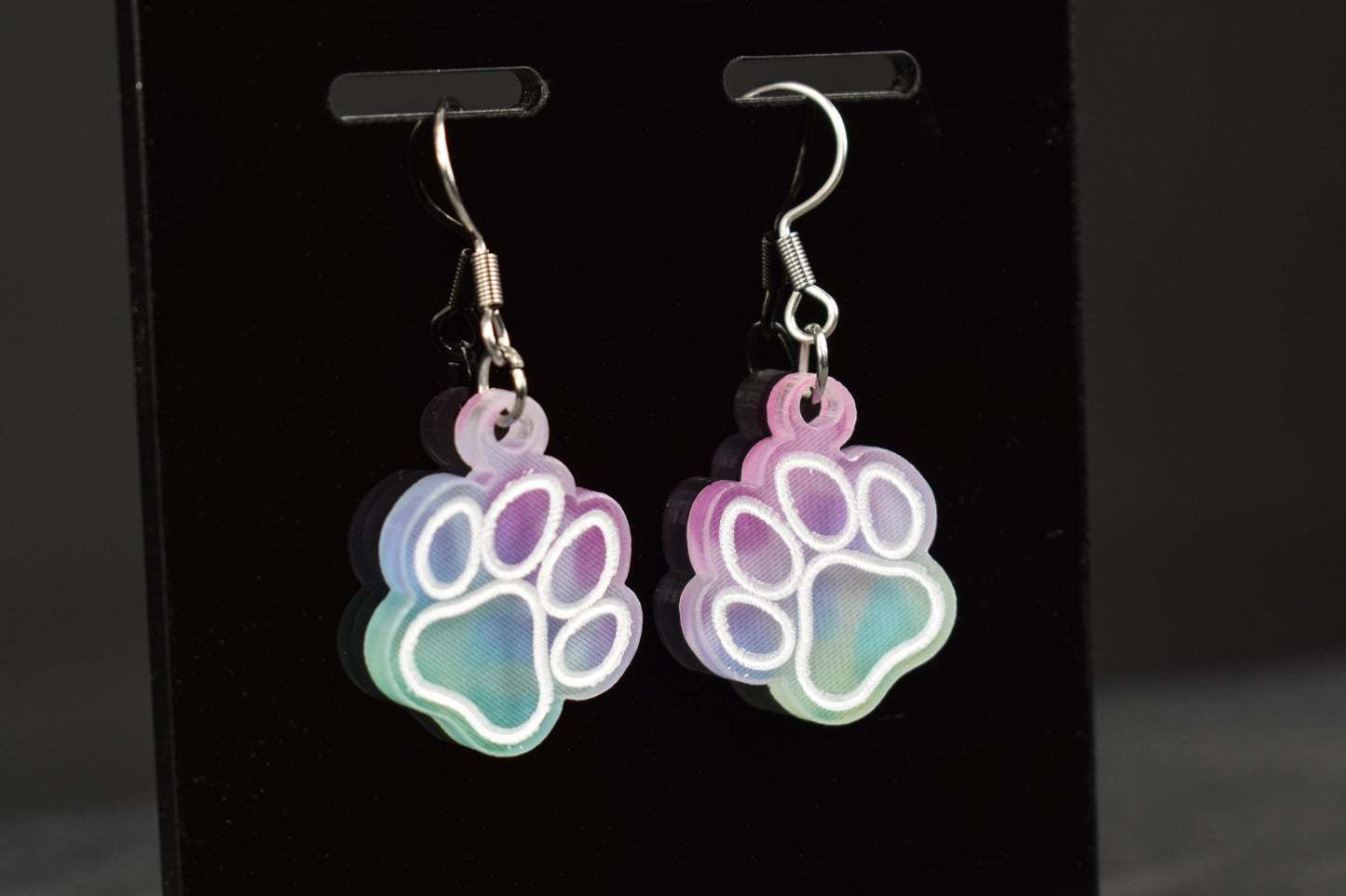 A close up of a pair of multicolored earrings with a paw print design and a cool hue, featuring silver hooks, displayed on a black stand