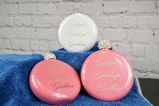 A trio of pink and white round glitter flasks with rhinestone caps, featuring a laser engraving of 'Gaslight Gatekeep Girlboss' set against a white brick background