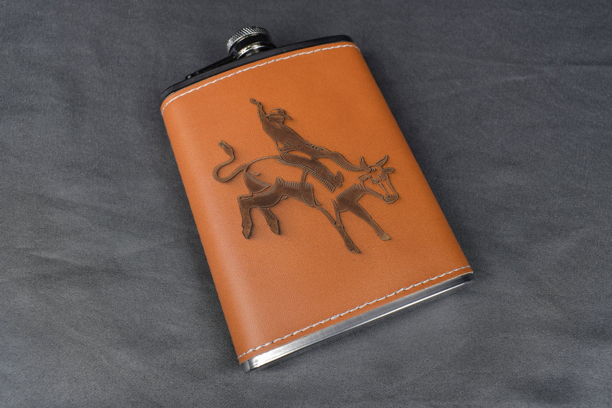 A brown leather wrapped metal flask with a bull rider design on the front sitting on a gray background