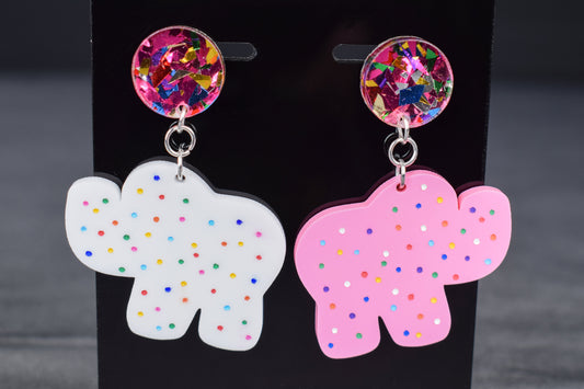 A pair of earrings featuring a white acrylic cookie-shaped elephant and a pink acrylic cookie-shaped camel, with a glittery multicolored acrylic circle attached to each and hand-painted dots