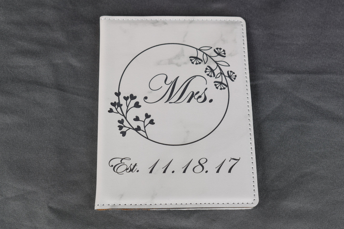 A white marble synthetic leatherette passport holder with 'Mrs.' and a date laser engraved on it, lying on a gray background