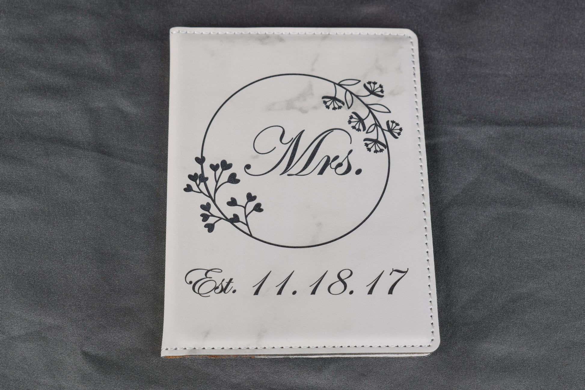 A white marble synthetic leatherette passport holder with 'Mrs.' and a date laser engraved on it, lying on a gray background