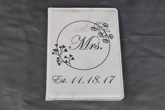 A white marble synthetic leatherette passport holder with 'Mrs.' and a date laser engraved on it, lying on a gray background