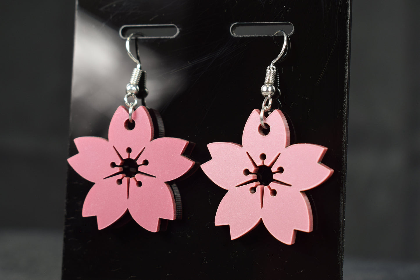 A pair of mauve metallic cherry blossom earrings with silver hooks, displayed on a black stand against a blurred background