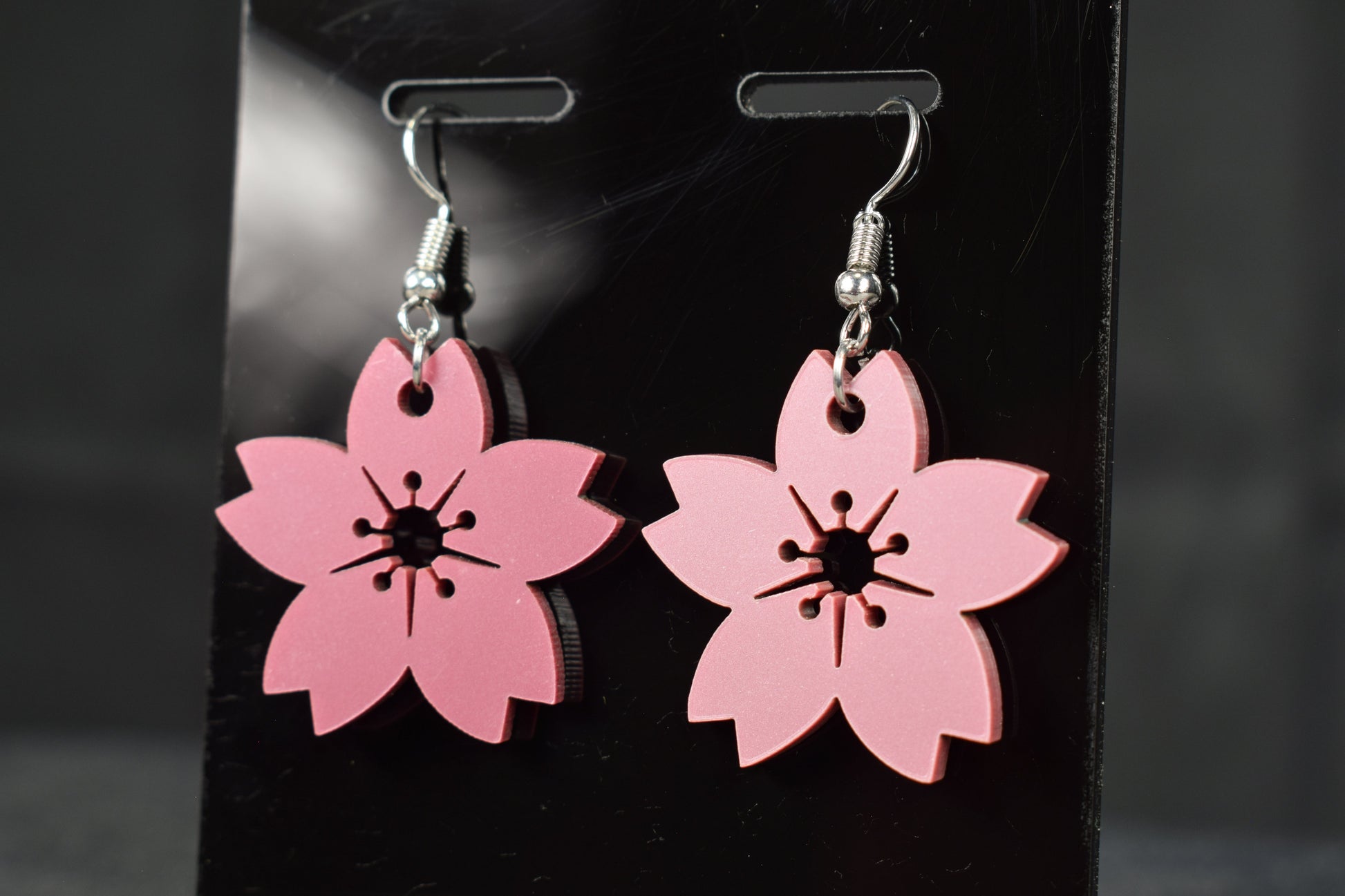 A pair of mauve metallic cherry blossom earrings with silver hooks, displayed on a black stand against a blurred background