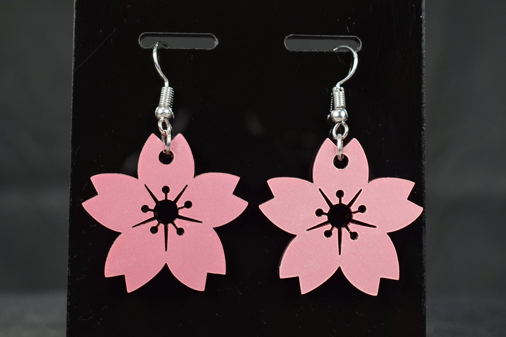 A pair of mauve metallic cherry blossom earrings with silver hooks, displayed on a black stand against a blurred background