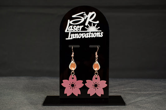 A pair of cherry blossom drop earrings in mauve metallic and iridescent colors, displayed on a black stand with the brand name 'SR Laser Innovations'.