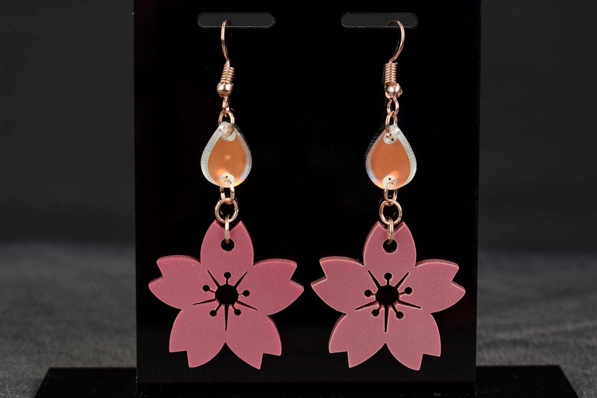 A pair of mauve metallic two-tier flower shaped earrings with iridescent accent teardrops displayed on a black stand, with rose gold hooks