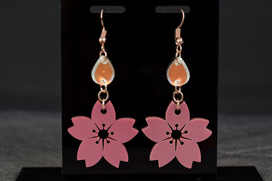 A pair of mauve metallic two-tier flower shaped earrings with iridescent accent teardrops displayed on a black stand, with rose gold hooks