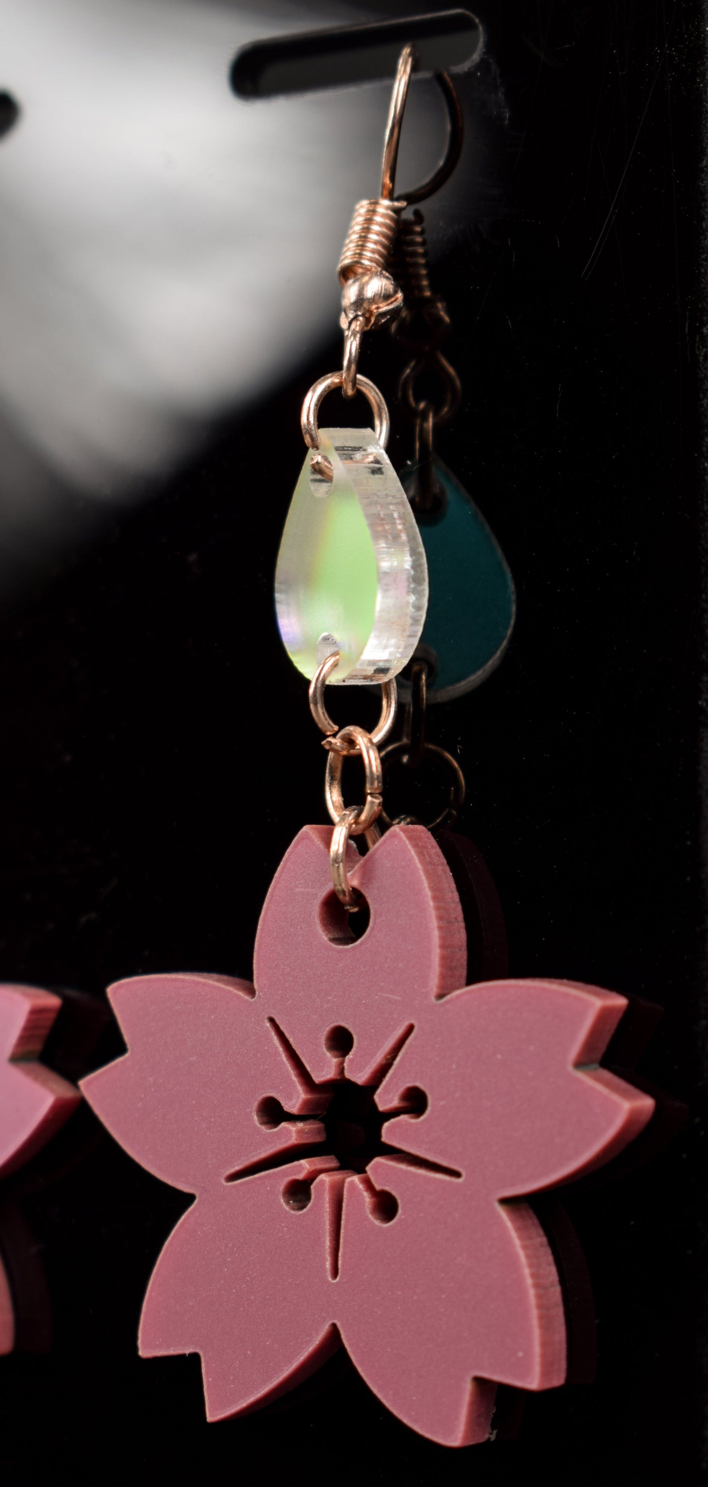 A mauve metallic two-tier flower shaped earring with an iridescent accent teardrop displayed on a black stand, featuring rose gold hooks
