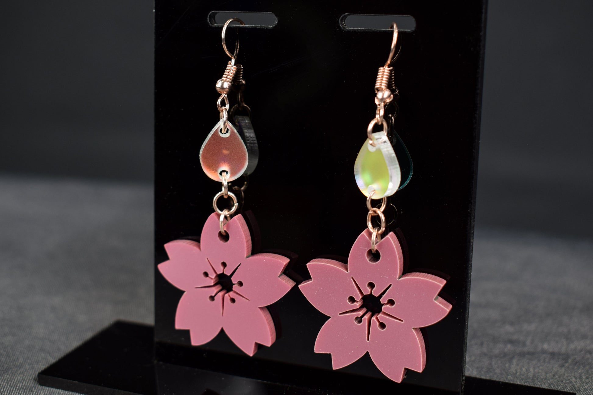 A pair of mauve metallic two-tier flower shaped earrings with iridescent accent teardrops displayed on a black stand with gold hooks
