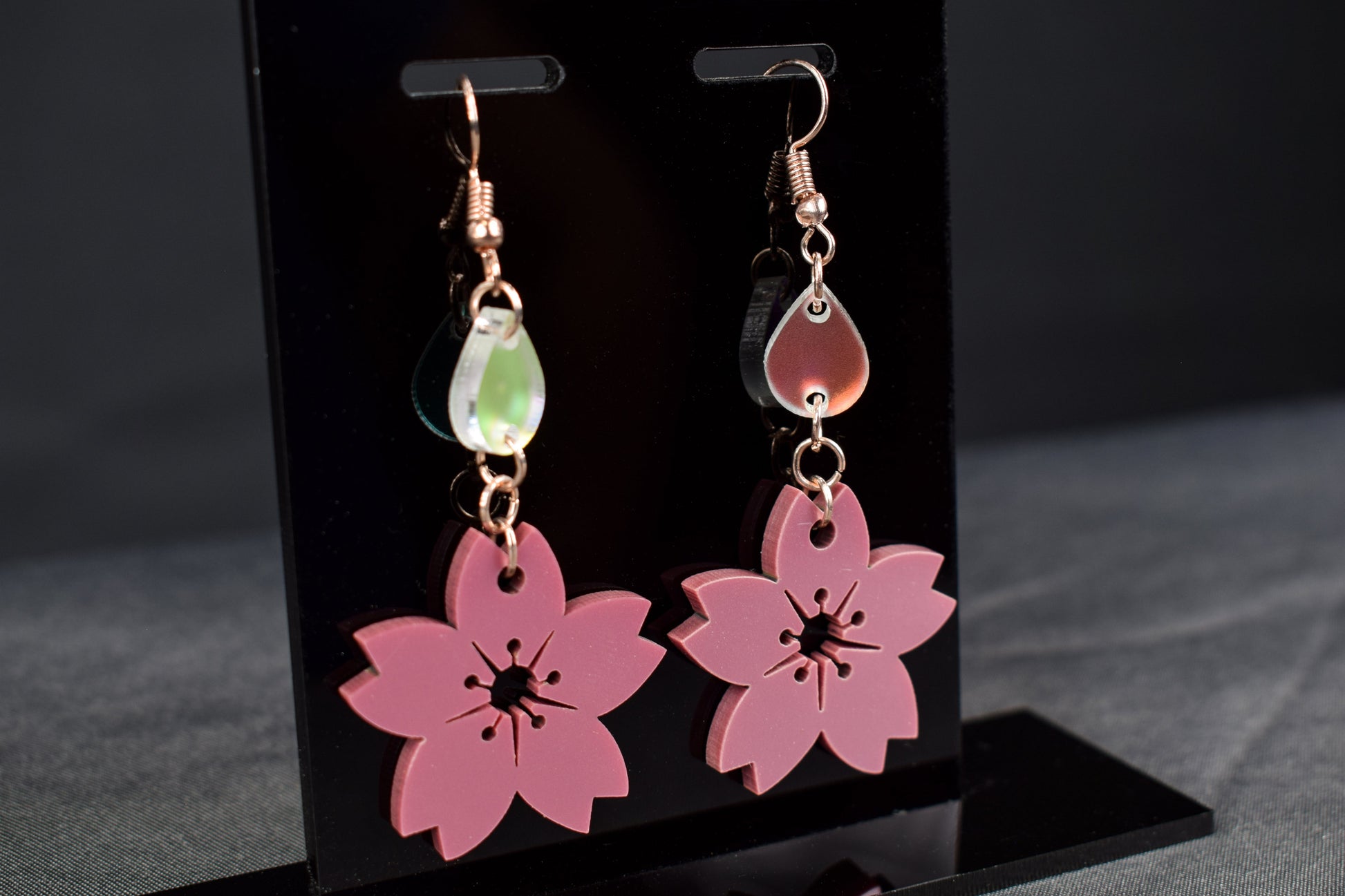 A pair of mauve metallic two-tier flower shaped earrings with iridescent accent teardrops displayed on a black stand