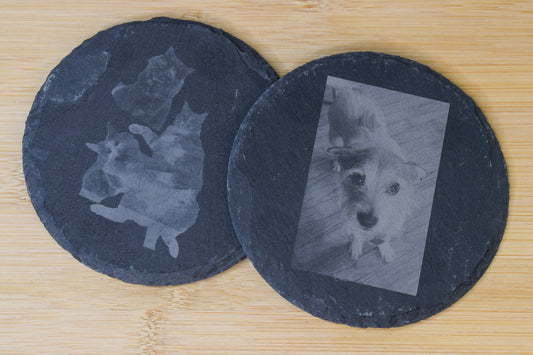 An overhead view of two circular slate coasters with images of cats and dogs engraved on top, laying on a wooden surface