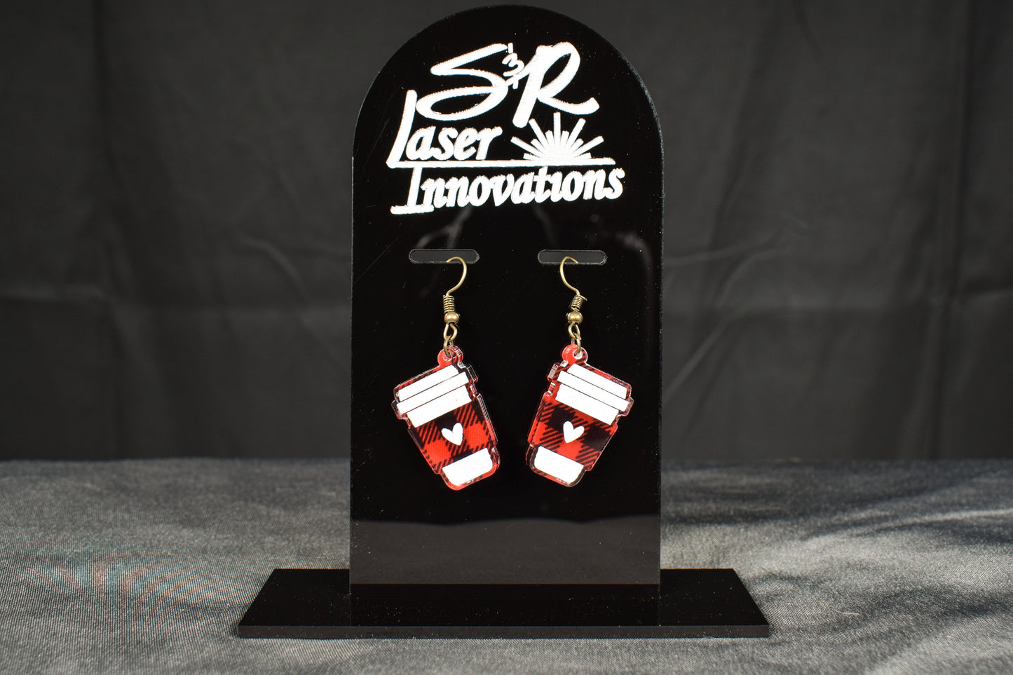 A pair of black and red plaid acrylic earrings with a coffee cup design, displayed on a black stand with the 'S&R Laser Innovations' logo 