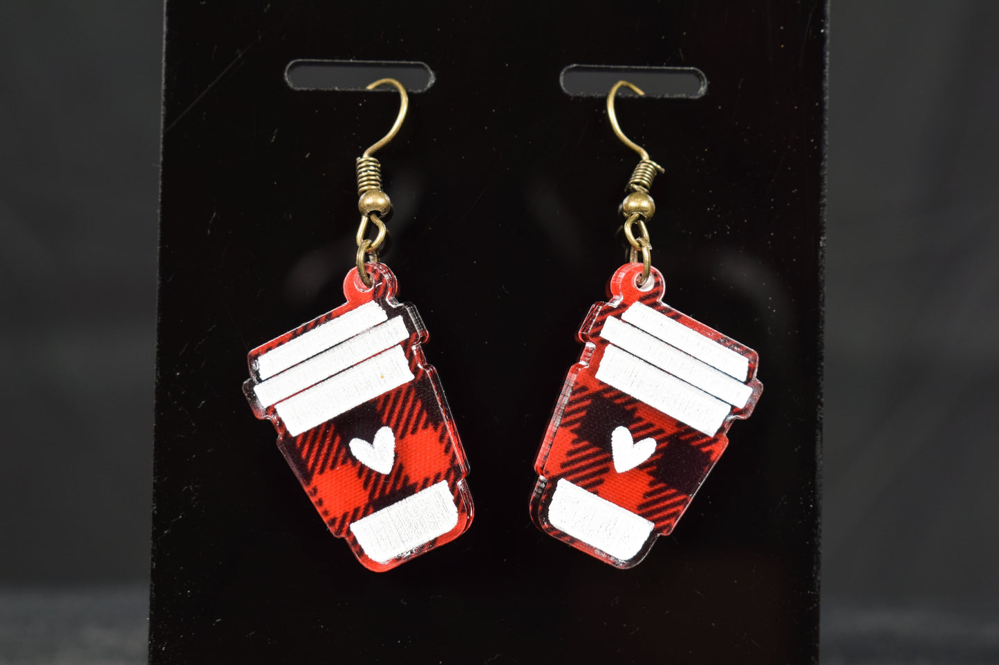 A pair of black and red plaid acrylic earrings with a coffee cup design and bronze hooks, displayed on a black stand