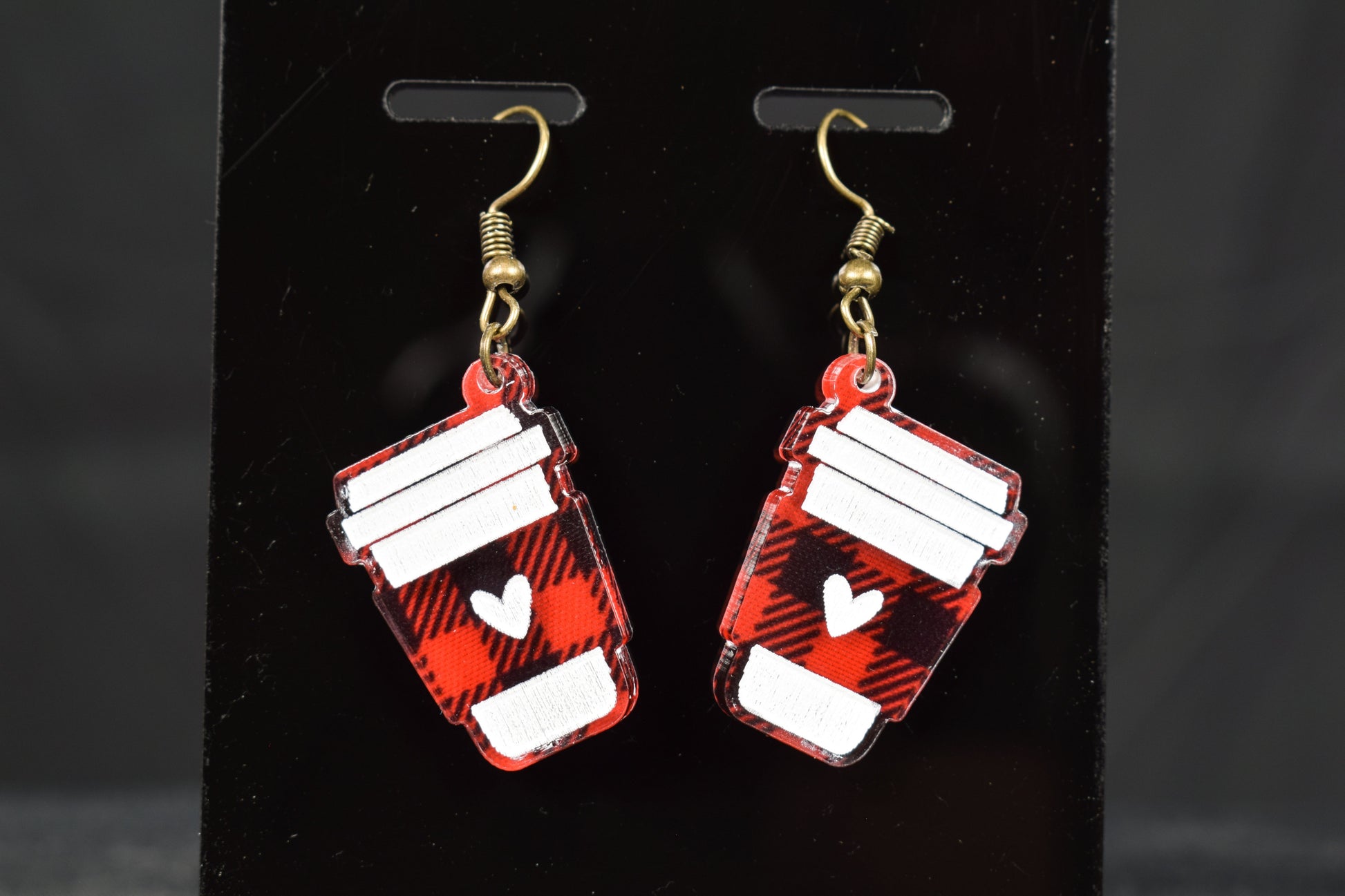 A pair of black and red plaid acrylic earrings with a coffee cup design and bronze hooks, displayed on a black stand