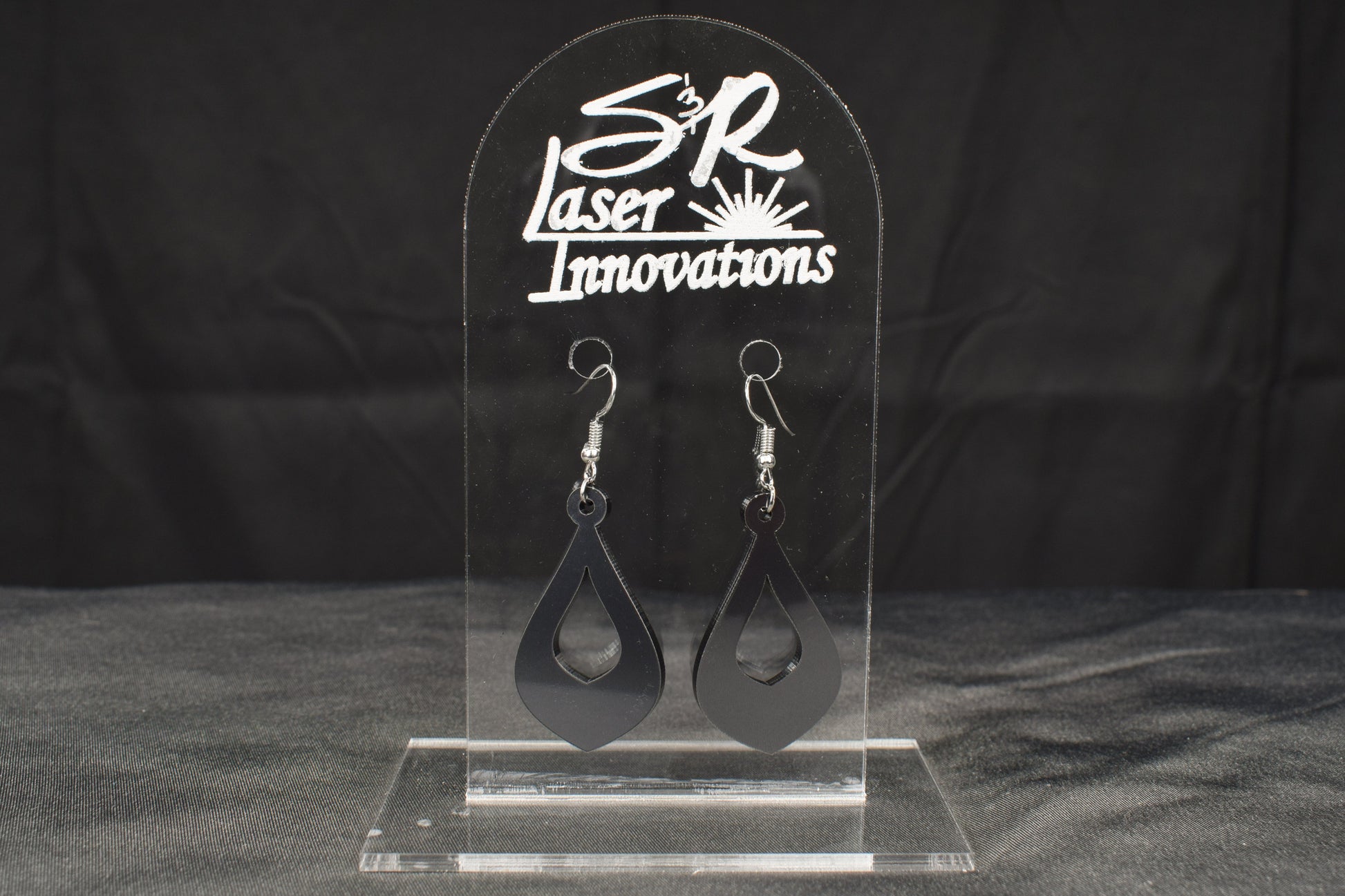 a pair of teardrop shaped hoop style earrings in charcoal metallic acrylic displayed on a clear stand with the 'S&R Laser Innovations' logo on top