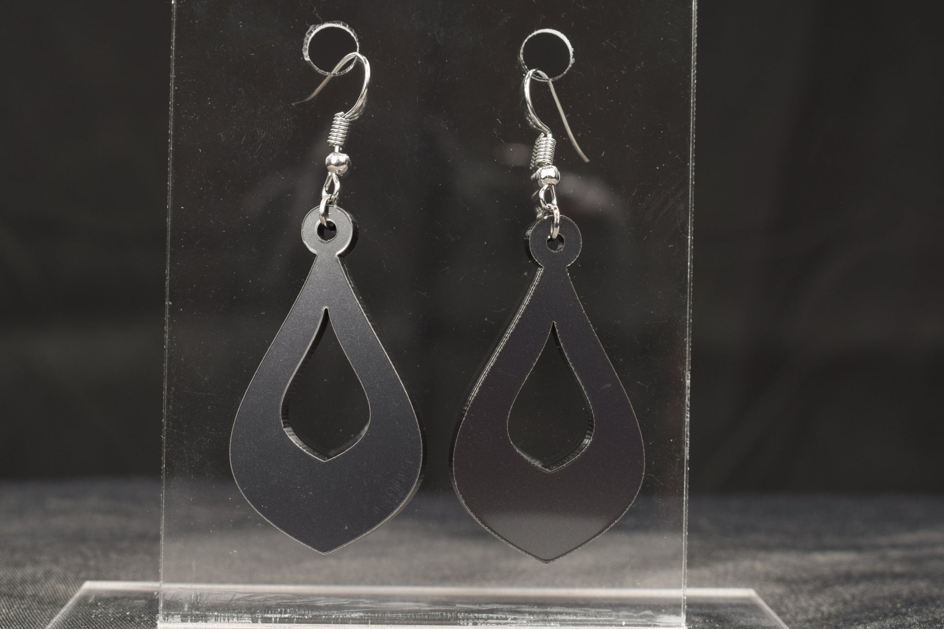 a pair of teardrop shaped hoop style earrings in charcoal metallic acrylic displayed on a clear stand, featuring stainless steel hooks