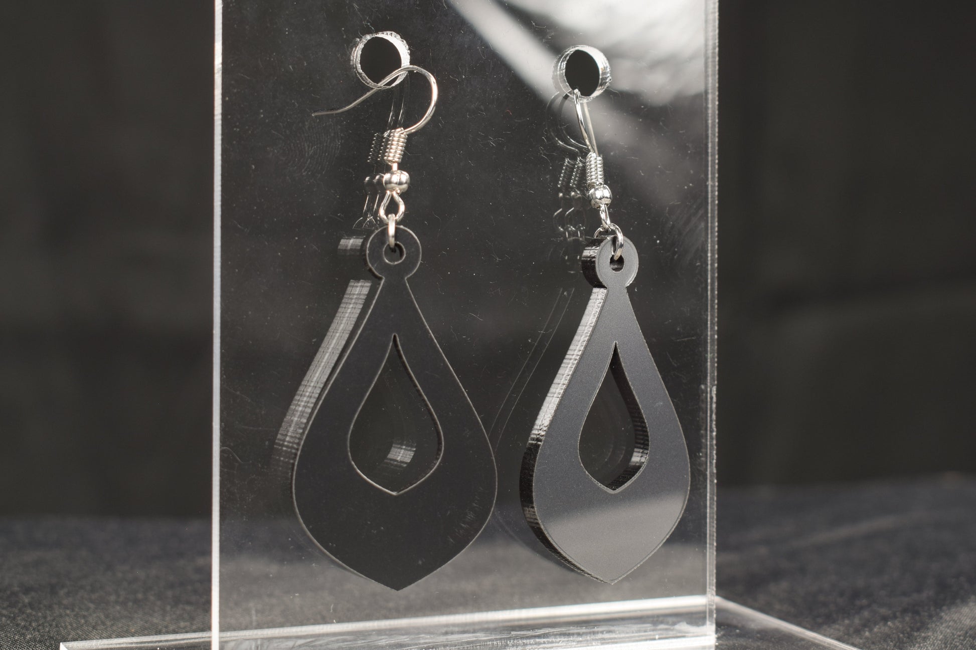A close up view of a pair of teardrop shaped hoop style earrings in charcoal metallic acrylic displayed on a clear stand 