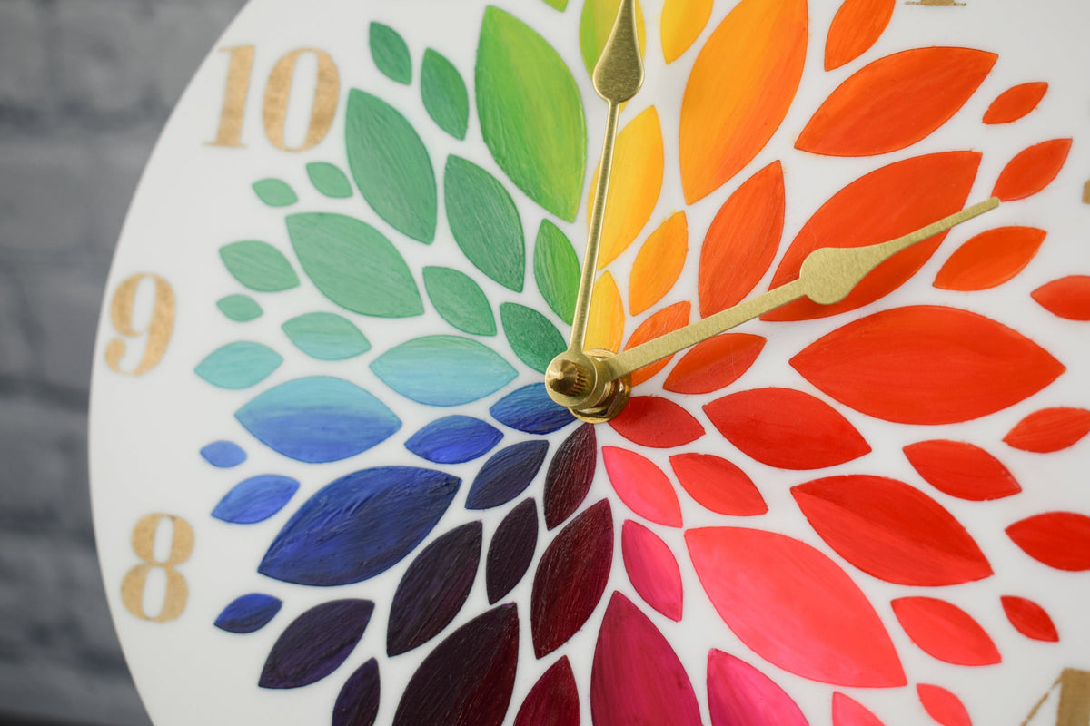 A close up view of a hand painted clock, showcasing the rainbow colored flower petals and gold hands, against a blurred background