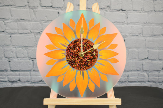 A hand-painted sunflower clock with glitter accents in the center, displayed on a wooden easel against a white brick background.