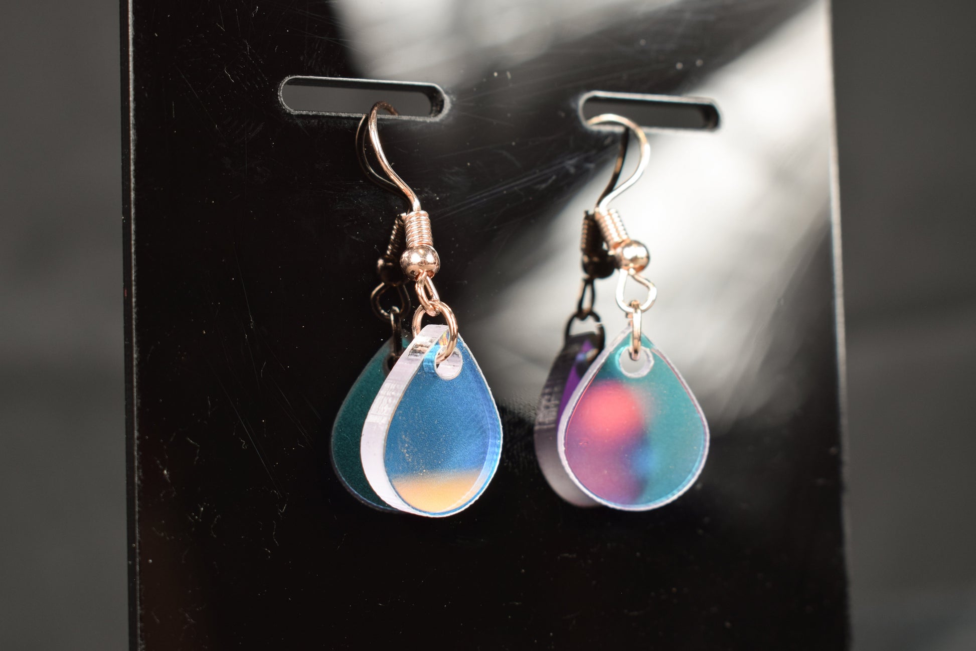 A close up view of a pair of iridescent rainbow teardrop earrings displayed on a black stand