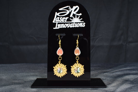 A pair of gold metallic and iridescent acrylic sunflower drop earrings displayed on a black stand with 'SR Laser Innovations' branding.