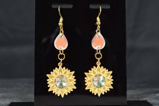 A pair of gold metallic and iridescent acrylic sunflower drop earrings displayed on a black stand