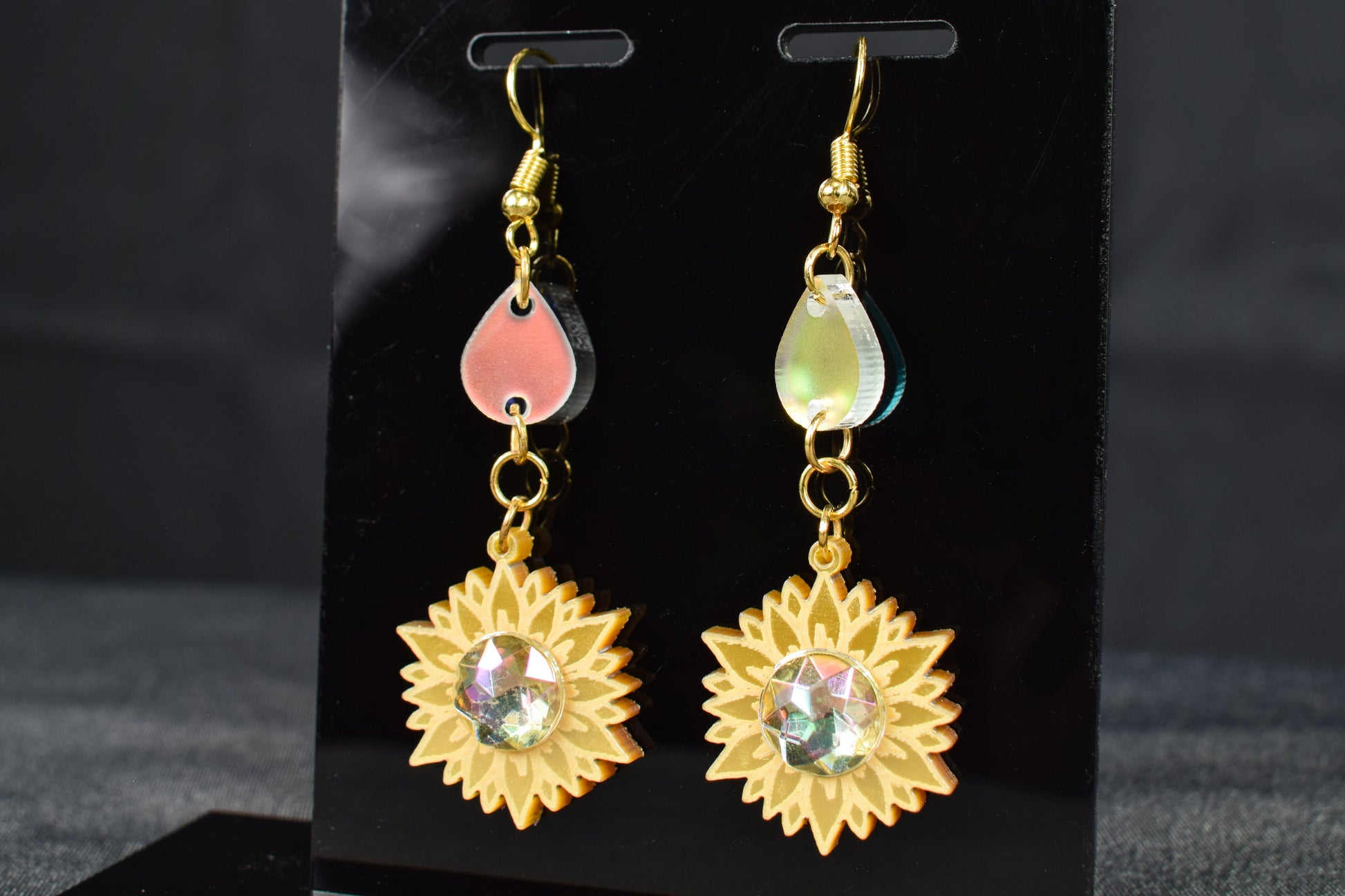 A pair of gold metallic and iridescent acrylic sunflower drop earrings displayed on a black stand