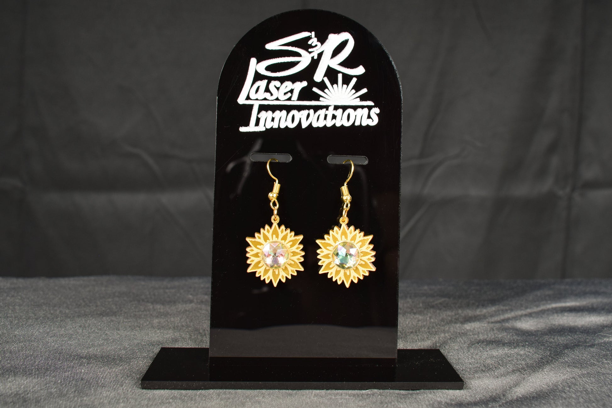 A pair of gold metallic acrylic earrings designed to resemble sunflowers with iridescent rhinestone centers, displayed on a black stand with 'SR Laser Innovations' branding.