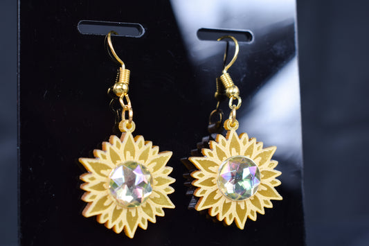 A close up of a pair of gold metallic acrylic earrings designed to resemble sunflowers with iridescent rhinestone centers, displayed on a black stand