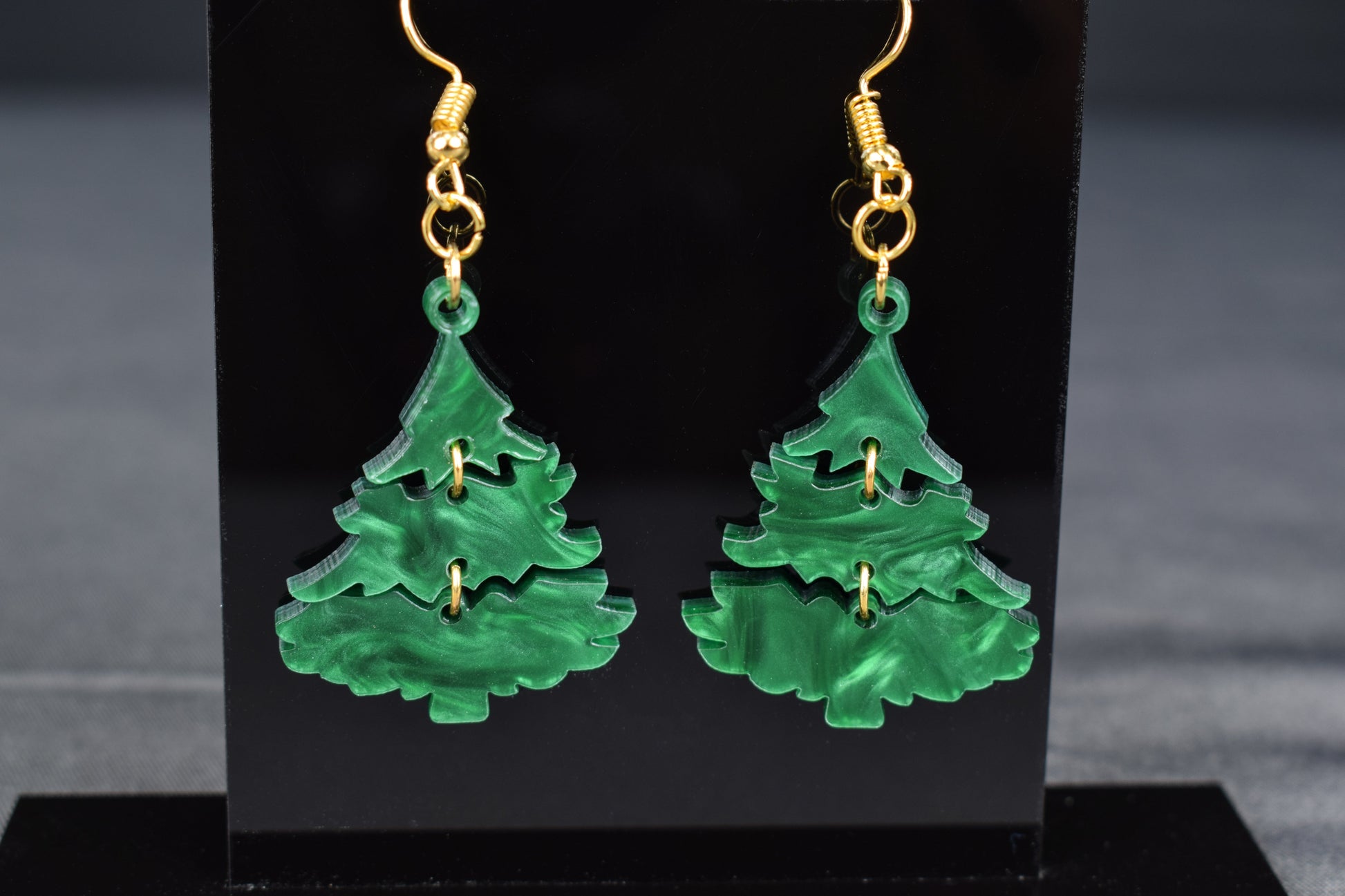 A pair of three-tiered Christmas tree earrings made of dark green swirl acrylic, displayed on a black stand with 'SR Laser Innovations' branding.