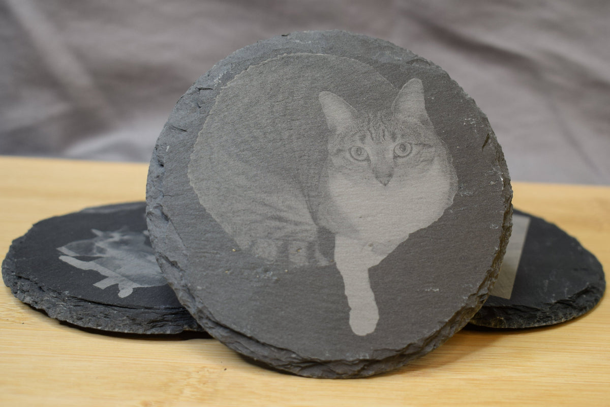 A round slate coaster with an engraved image of a cat in the center, displayed on a wooden surface.