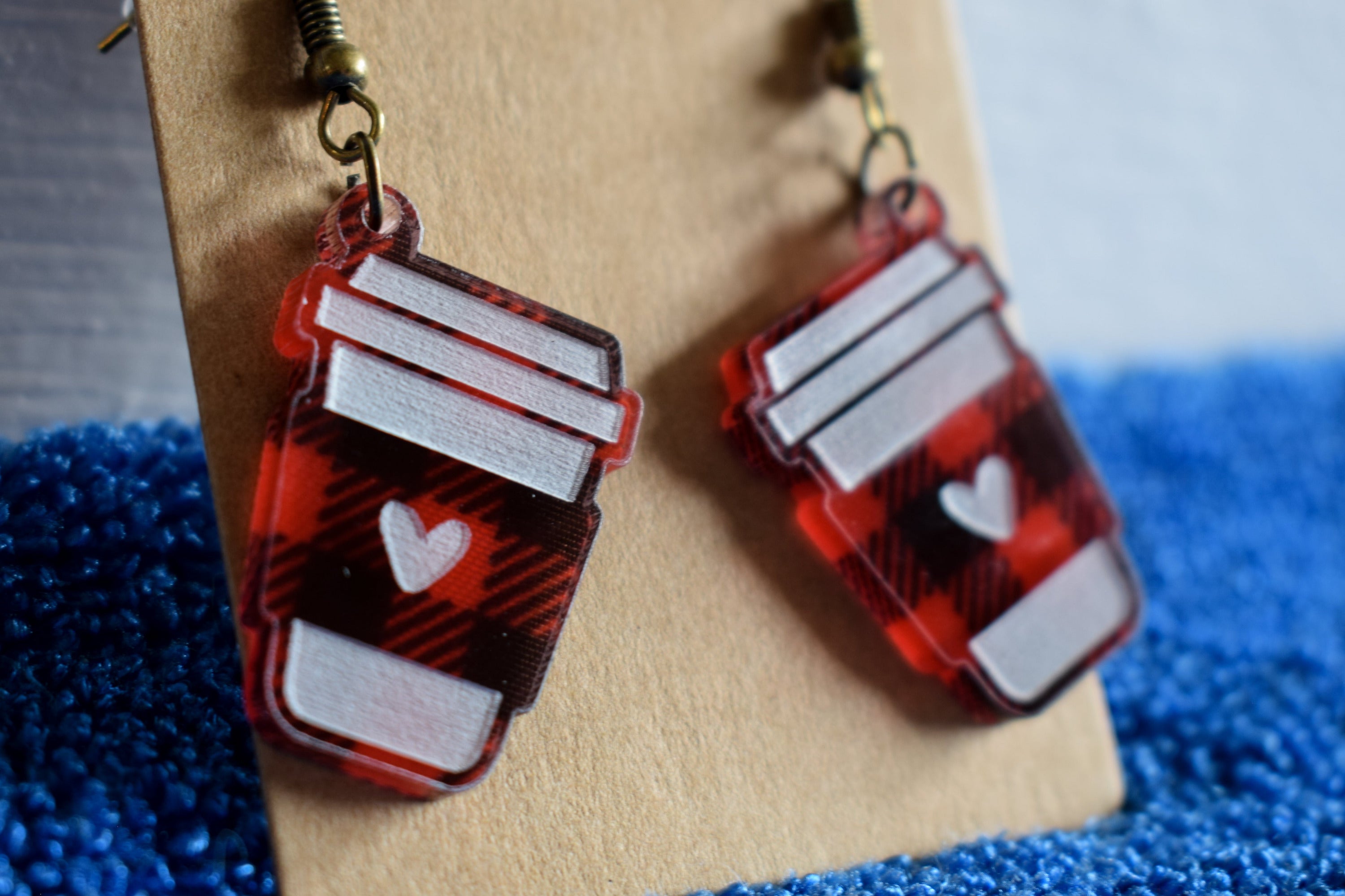 A pair of black and red plaid acrylic earrings with a coffee cup design and bronze hooks, displayed on a brown card with a blurred blue and white background