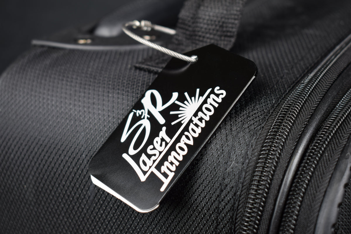 A black aluminum luggage tag with custom logo engraving, attached to a piece of luggage.
