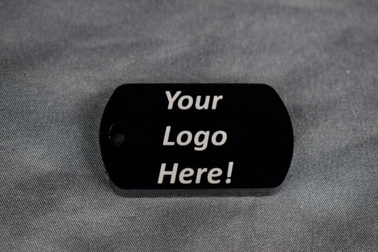 a black acrylic dogtag shaped keychain with the text 'your logo here' laser engraved on top, laying on a gray surface