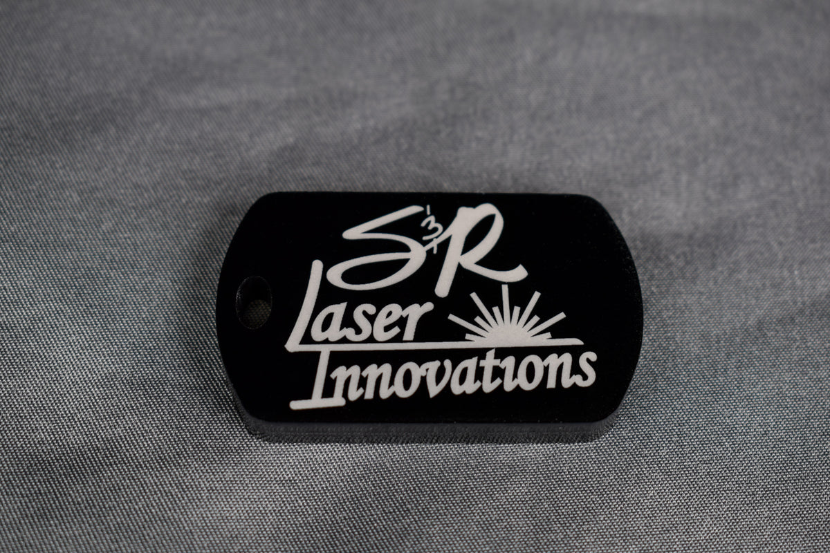 A black acrylic dogtag-style keychain with white 'S&R Laser Innovations' logo engraving on the front side, lying on a textured grey surface.