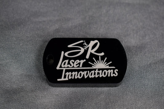 A black acrylic dogtag-style keychain with white 'S&R Laser Innovations' logo engraving on the front side, lying on a textured grey surface.