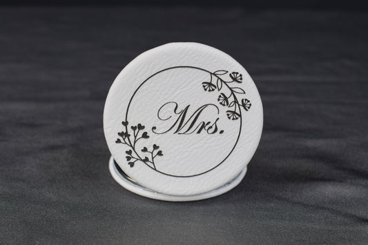 A round compact mirror with a white synthetic leatherette cover, featuring a laser-engraved design with the text 'Mrs.' and floral elements.