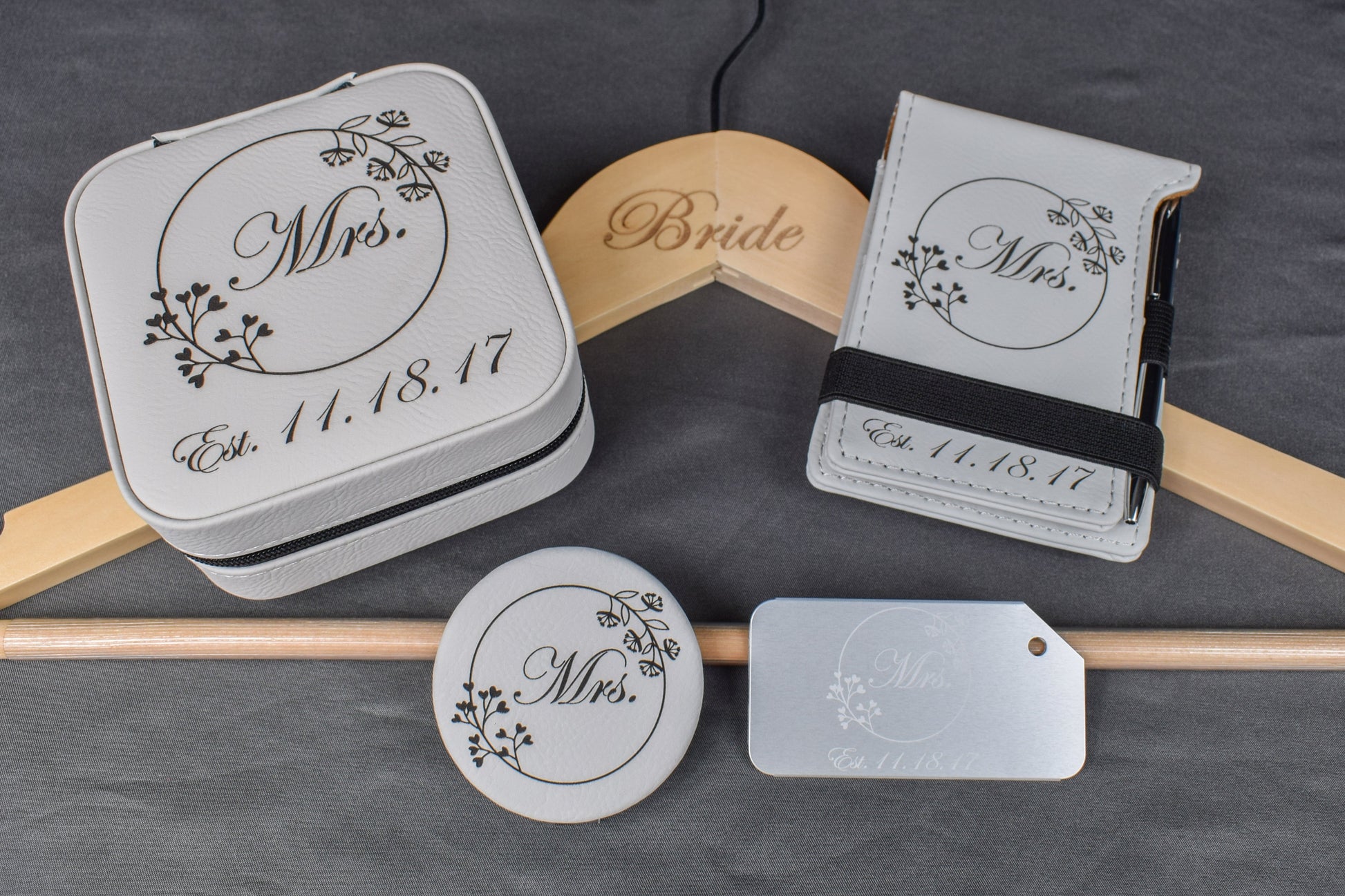 A set of personalized bridal gift items including a custom aluminum luggage tag, a white compact mirror, a white vow mini notebook, a custom engraved bridal hanger, and a custom laser engraved travel jewelry box, all with elegant designs and the wedding date.