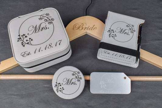 A set of personalized bridal gift items including a custom aluminum luggage tag, a white compact mirror, a white vow mini notebook, a custom engraved bridal hanger, and a custom laser engraved travel jewelry box, all with elegant designs and the wedding date.