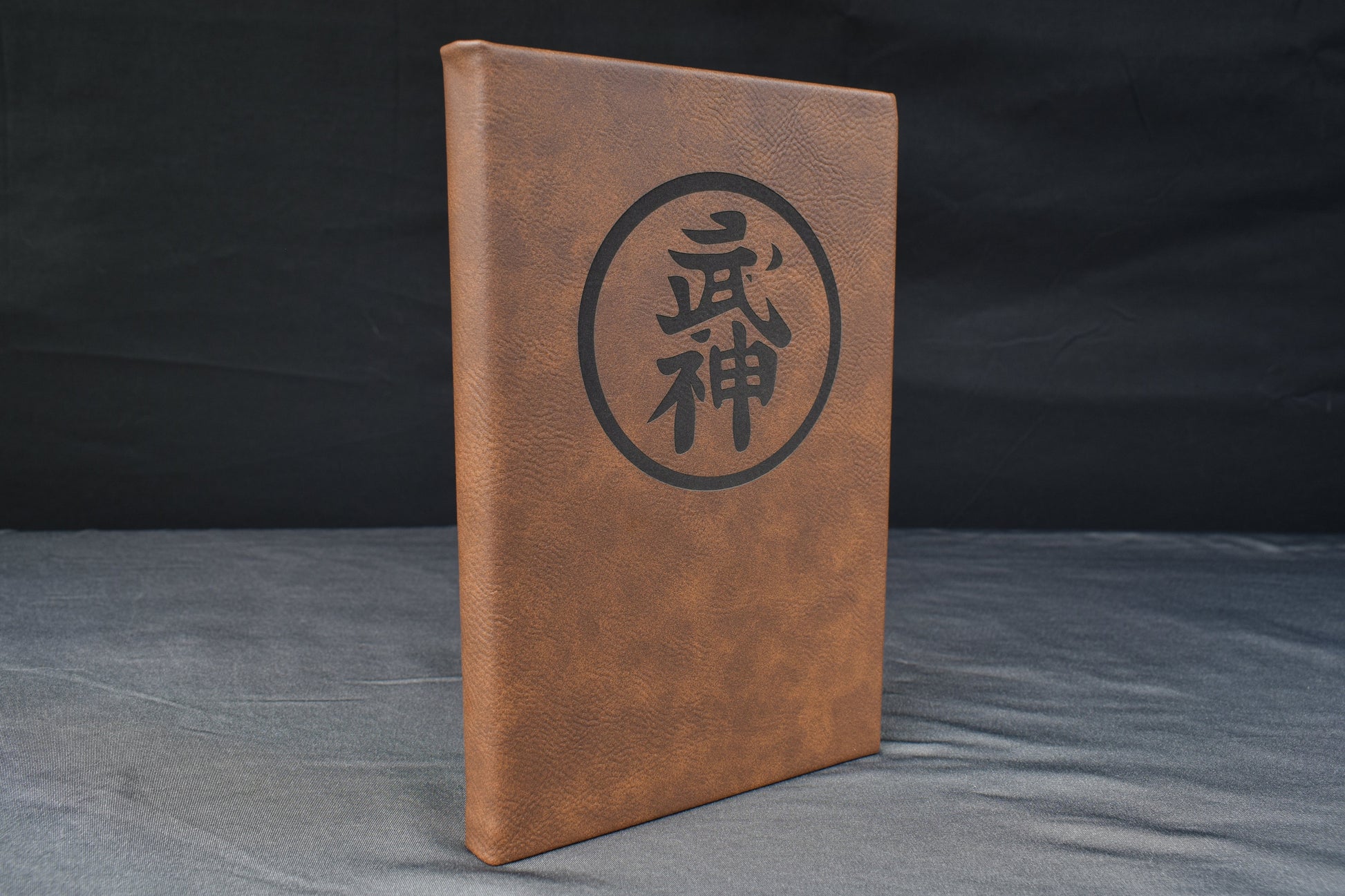a brown leatherette journal with a prominent symbol laser engraved on the cover, against a black background