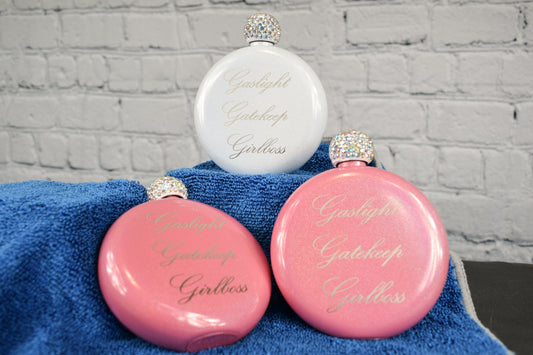 A trio of pink and white round glitter flasks with rhinestone caps, featuring a laser engraving of 'Gaslight Gatekeep Girlboss' set against a white brick background