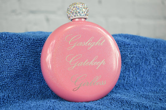 A pink glittery flask with a white rhinestone top, engraved with the phrase 'Gaslight Gatekeep Girlboss' on a blue towel.