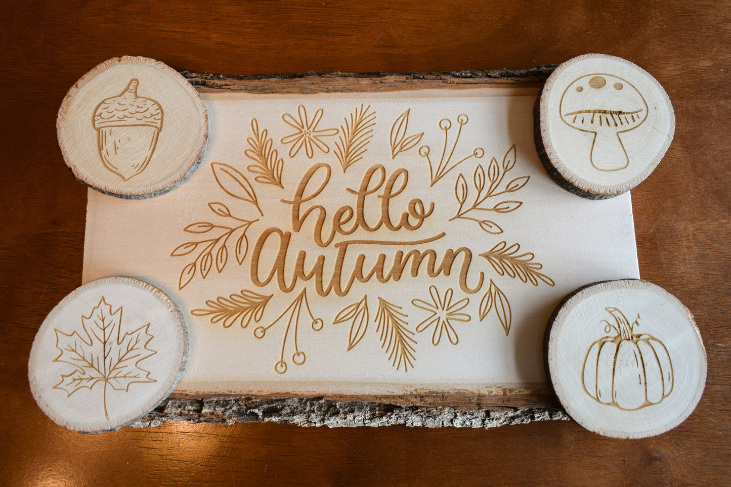 A set of Autumn-themed charcuterie board and coasters. The board has 'Hello Autumn' text and leaf, pumpkin, and acorn designs engraved on it, and is presented with four coasters, each with a different fall-themed engraving.