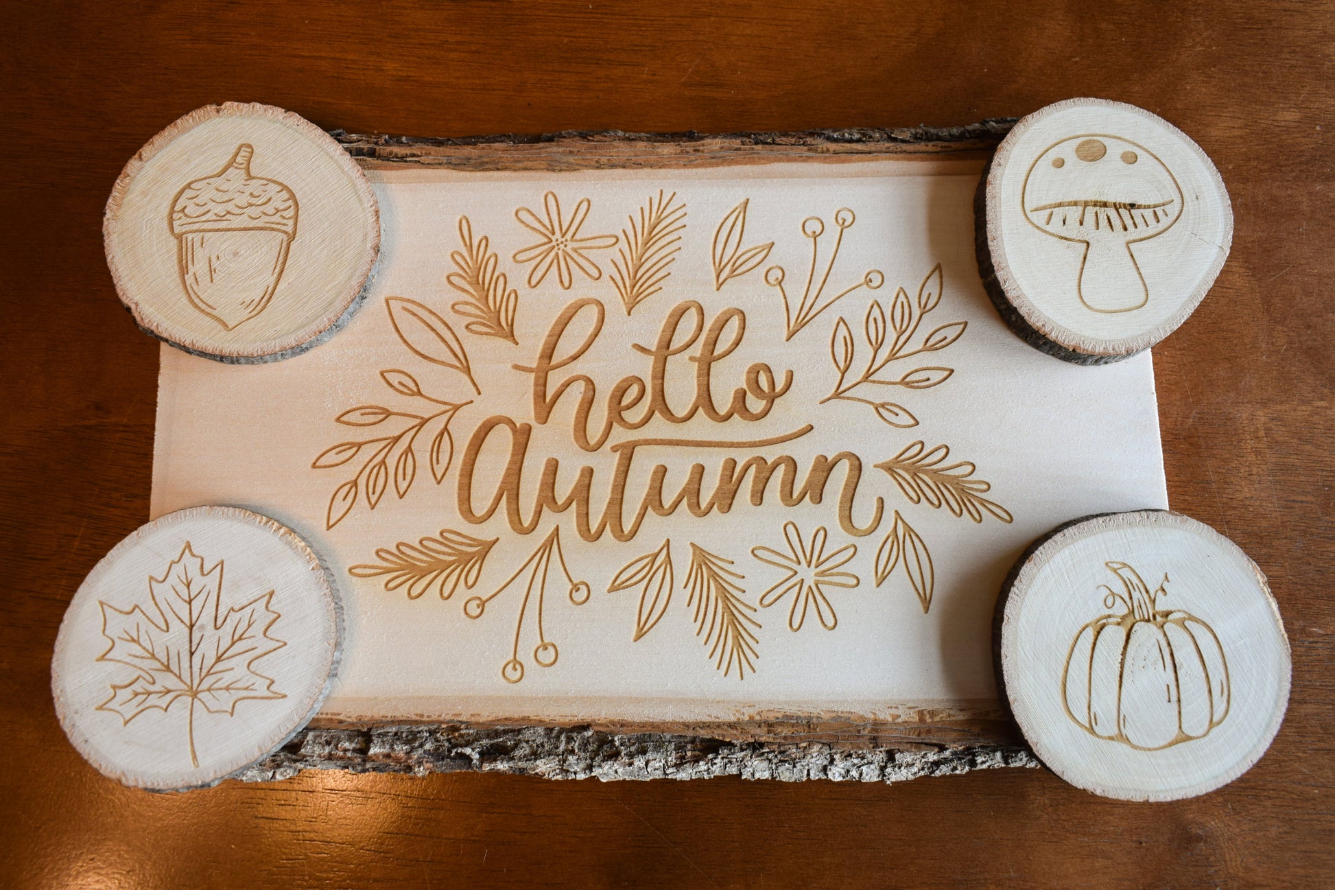 A set of Autumn-themed charcuterie board and coasters. The board has 'Hello Autumn' text and leaf, pumpkin, and acorn designs engraved on it, and is presented with four coasters, each with a different fall-themed engraving.