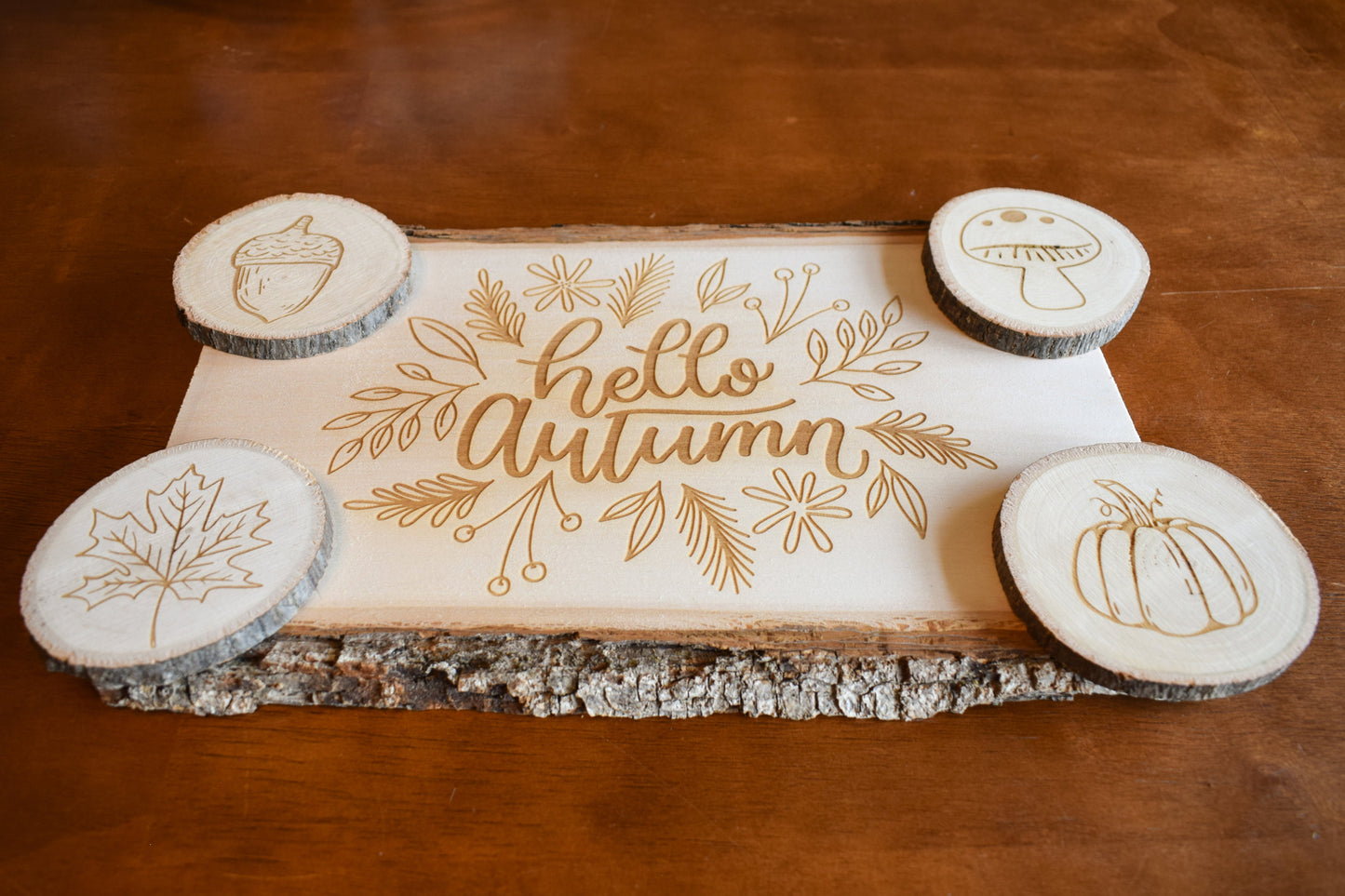 An angled view of a set of Autumn-themed charcuterie board and coasters. The board has 'Hello Autumn' text and leaf, pumpkin, and acorn designs engraved on it, and is presented with four coasters, each with a different fall-themed engraving.