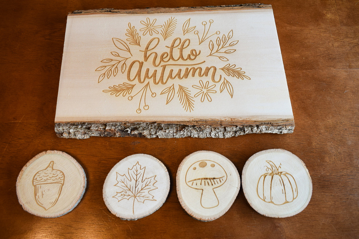 A set of Autumn-themed charcuterie board and coasters. The board has 'Hello Autumn' text and leaf, pumpkin, and acorn designs engraved on it, and is presented with four coasters below it, each with a different fall-themed engraving.