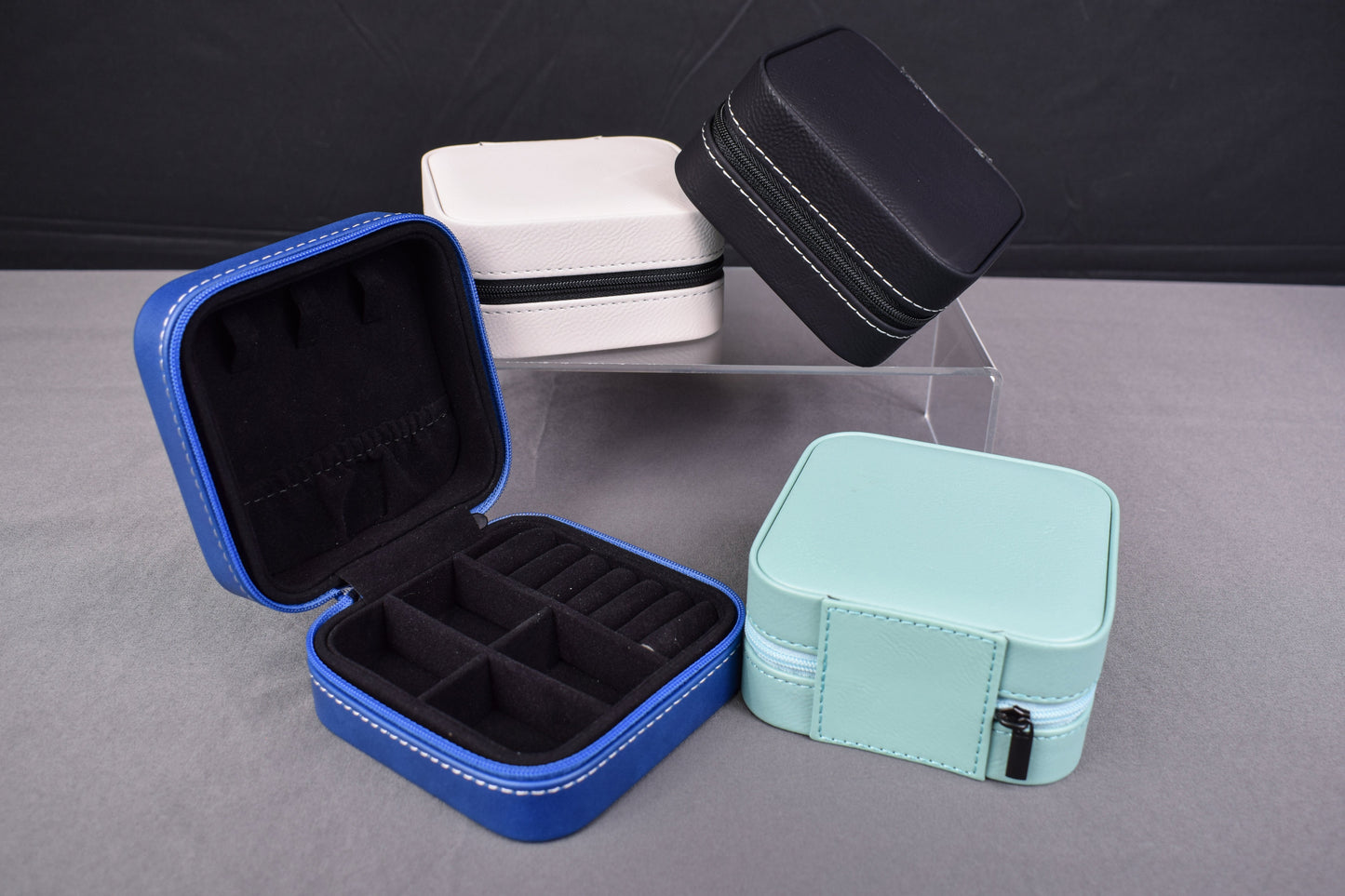 A collection of synthetic leatherette travel jewelry boxes in blue, black, white, and teal colors, displayed open to show the interior compartments, on a gray surface.